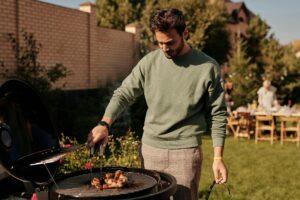 A man grills chicken outdoors at a sunny backyard barbecue gathering.