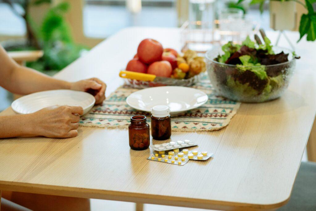 From above of crop anonymous person sitting at table with pile of various medicines and bowls of fresh lettuce salad and ripe fruits in kitchen