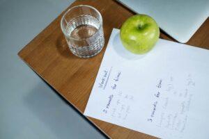 A green apple, glass of water, and workout plan on a wooden desk symbolize healthy living.