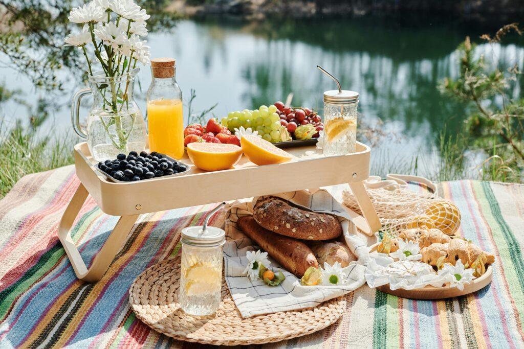 A colorful picnic setup with fresh fruits, bread, and drinks by a serene lake.